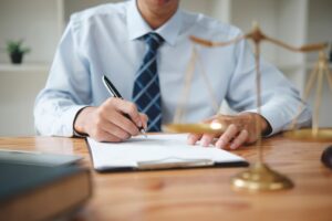 A lawyer works on a claim for psychological injury. He is sat at a desk filling out paperwork while wearing a suit. There is a scales of justice in the foreground