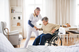 A health care worker helping an elderly woman who is a care home resident get out of a wheelchair and into her bed.