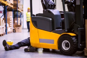 A yellow forklift next to a warehouse worker lying on the ground